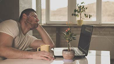 Guy fell asleep at a desk with a laptop and with a cup of tea at the white table in the kitchen