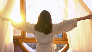 A young woman in a white coat walks to the window and opens the curtains. The girl in the morning looks out the window