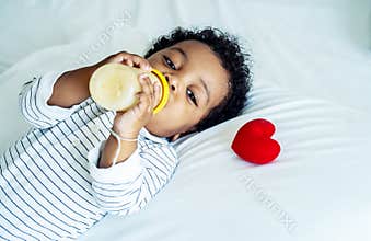 African american cute baby boy drinking milk from bottle and sleeping on bed with a red heart at home, Children need love