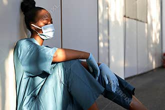 Tired exhausted woman african nurse wear face mask gloves sit on hospital floor.