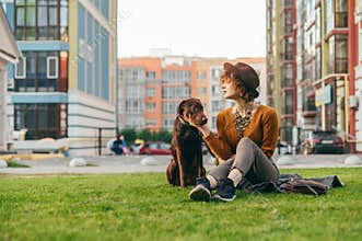 Portrait of a stylish girl in a hat sits with a beautiful brown dog on the lawn against the backdrop of a modern apartment