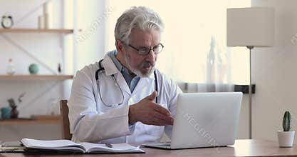 Happy older hoary male doctor waving hello to distant client.