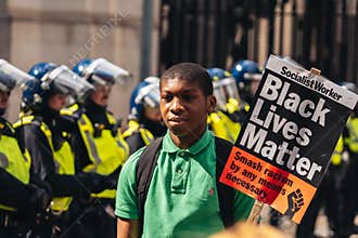 Black Lives Matter protest during lockdown coronavirus pandemic. Young man with BLM banner posing in front of Police officers