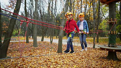 Two little girls in orange helmets and protective gear on rope-way in autumn  forest. Children are engaged climbing rope park.