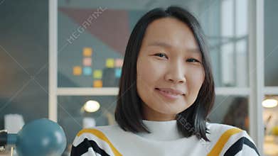 Portrait of charming Asian girl looking at camera then smiling indoors in office