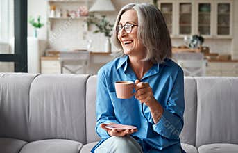 Happy relaxed mature older woman drinking coffee relaxing on sofa at home.