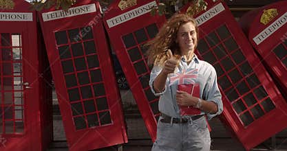 Smiling woman showing thumbs up, standing against British telephone, studying English.