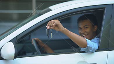 Happy teenager showing car key to window sitting on driver seat of new vehicle