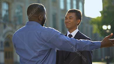 Glad parent hugging young son in suit outdoors university, prom celebration