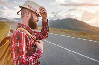 Portrait of a bearded happy serious traveler hipster with a backpack in a plaid shirt and a hat next to an unknown car