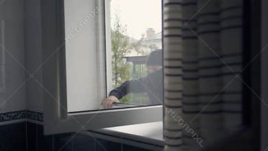 Close-up of the thief in black clothes and balaclava breaking into the house through the window. Concept of offense