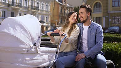 Cheerful husband wife hugging swinging newborn in stroller, parenthood happiness
