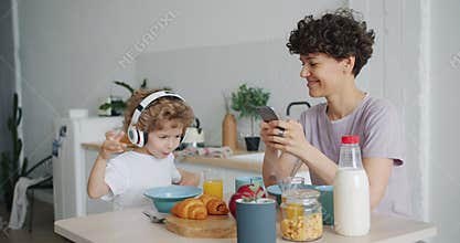 Young lady using smartphone while small boy enjoying music at kitchen table