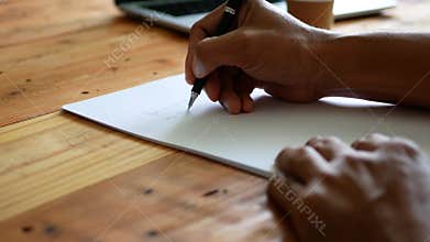 A man`s hand writing on a white paper on a table with a cup of coffee and a computer notebook nearby