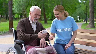 Female volunteer helping disabled old man use smartphone, nursing home charity
