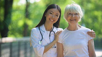 Nurse hugging happy senior woman and smiling into camera, day in hospital park
