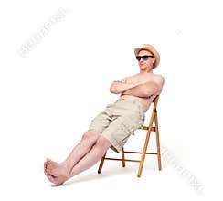 Happy relaxed man in shorts, hat and sunglasses resting on a chair under the sun,  on white background