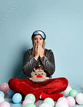 Teenage girl in reindeer pajamas, red socks, sleep mask. Looking surprised, sitting on floor, balloons, blue background. Close up