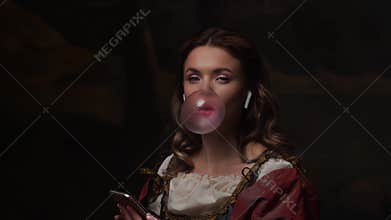 Medieval teen, young girl in vintage dress uses smartphone and blows bubble gum