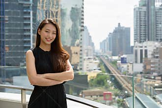 Happy young beautiful Asian businesswoman smiling with arms crossed against view of the city