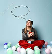 Teenager in reindeer pajamas, red socks, sleep mask. Holding cup, dreaming, sitting on floor, balloons, blue background. Close up