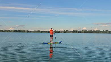 A man swims standing on a SUP board and rowing. A young man swims on a SUP board on a lake. Stand up paddle boarding