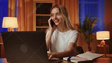 A smiling blonde student girl uses a laptop and talks on the smartphone within a fellow student about the upcoming exam