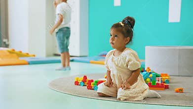 Little dark-skinned girl in dress sitting on carpet with toy plastic blocks looking around during playtime in nursery