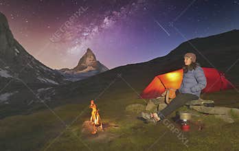Climber resting next to the tent and fire, while preparing a hot tea, Matterhorn peak behind and the Milky Way