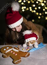 Girl with a cat in santa hats decorated a gingerbread.