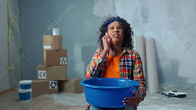 Upset African American woman is holding blue bowl into which drops of water are dripping from ceiling leak. Female is
