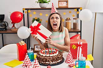 Hispanic woman celebrating birthday with cake holding gift angry and mad screaming frustrated and furious, shouting with anger