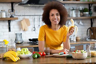 Healthy food concept. Healthy african american curly haired young woman stands at home in the kitchen, holds banana in her hand,