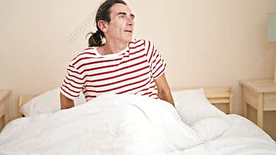 Middle age man waking up sitting on bed at bedroom