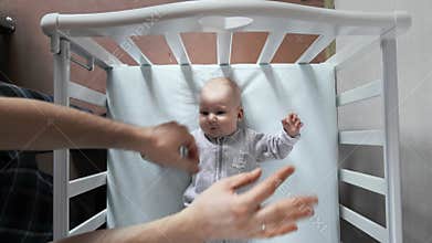 Father moves his arms to make his baby fall asleep. Two three month old infant is lying in a crib and his male parent