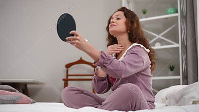 Confident mature woman applying moisturizer on neck skin smiling sitting on bed at home. Wide shot of Caucasian lady in