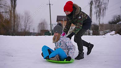 Cheerful teenage boy pushing sled with joyful girl sitting enjoying leisure. Wide shot side view happy carefree