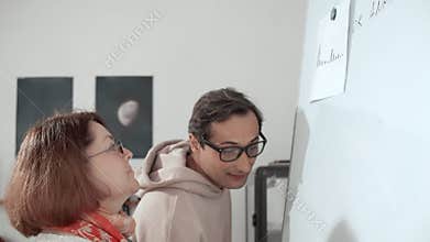 An adult plump woman with glasses comes up to a white board and a man with glasses explains to her and writes on the