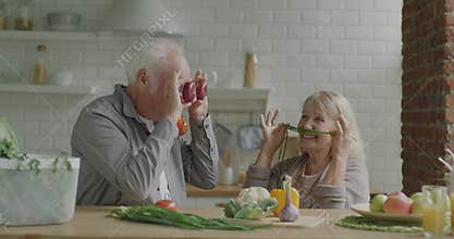 Portrait of joyful old people husband and wife having fun with vegetables making grimaces in kitchen at home