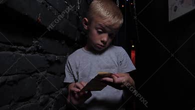 A little boy in a dark quest room, he unfolds an old folded note with a task.