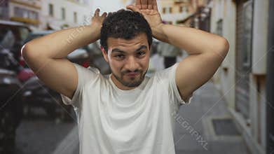 Man in white tshirt holds hands behind head on street with blurred buildings and parked scooters in view; playfulness amusement