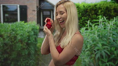 Blonde woman holds ringbox with hands and smiles in front of building with hedges behind; romance commitment