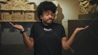 Man with afro shrugging while wearing staff uniform in museum indoor setting, standing against artifact display, suggests