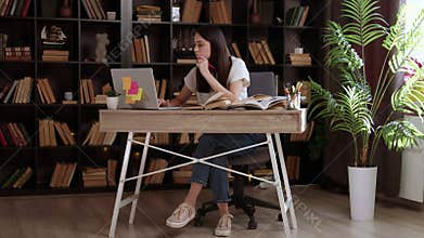 Curious and thoughtful young woman sitting in front of a computer searching for information on the internet and in books