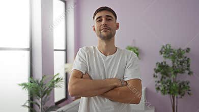 Young man in white shirt with arms crossed in bright living room setting with potted plants and large windows creating an airy and