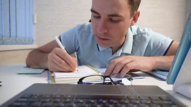 Young man with serious face using laptop computer, typing on keyboard, searching information online, writing notes
