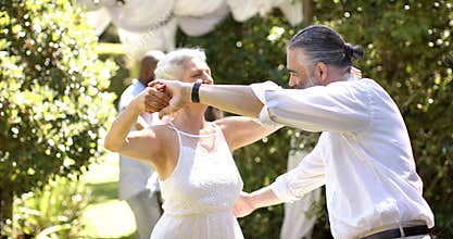 Dancing together, senior bride and groom enjoying outdoor wedding event with friends in background