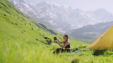 Woman on backpacking trip cooks outdoors with mini gas stove in mountain meadow, makes coffee in pot, turns off burner