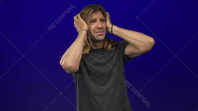 Blond man with long hair wearing dark grey shirt pressing hands over ears in bright blue studio; distress