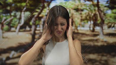 Woman presses fingers into bare ears in green park amid trees with sunlight filtering through leaves; frustration